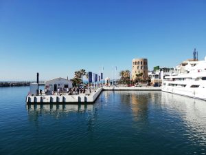 Tranquil sunny view of a harbor in Spain with pier, people, and yachts reflecting on the water.